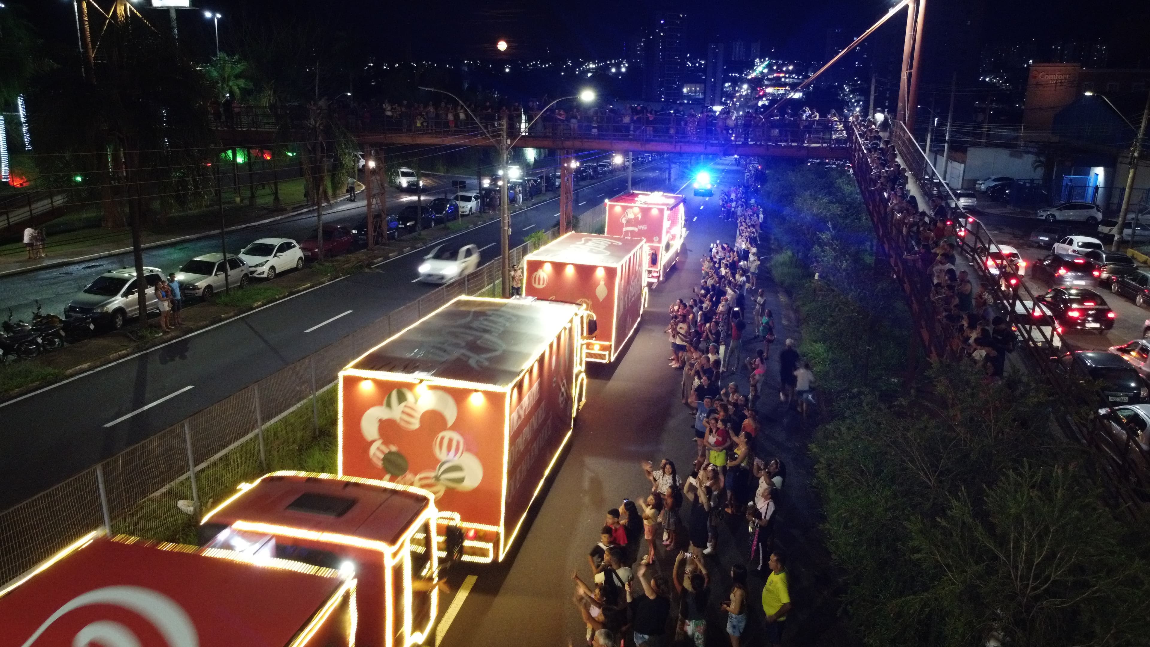 Caravana de caminhões iluminados de Natal à noite, com público na avenida, vista de cima